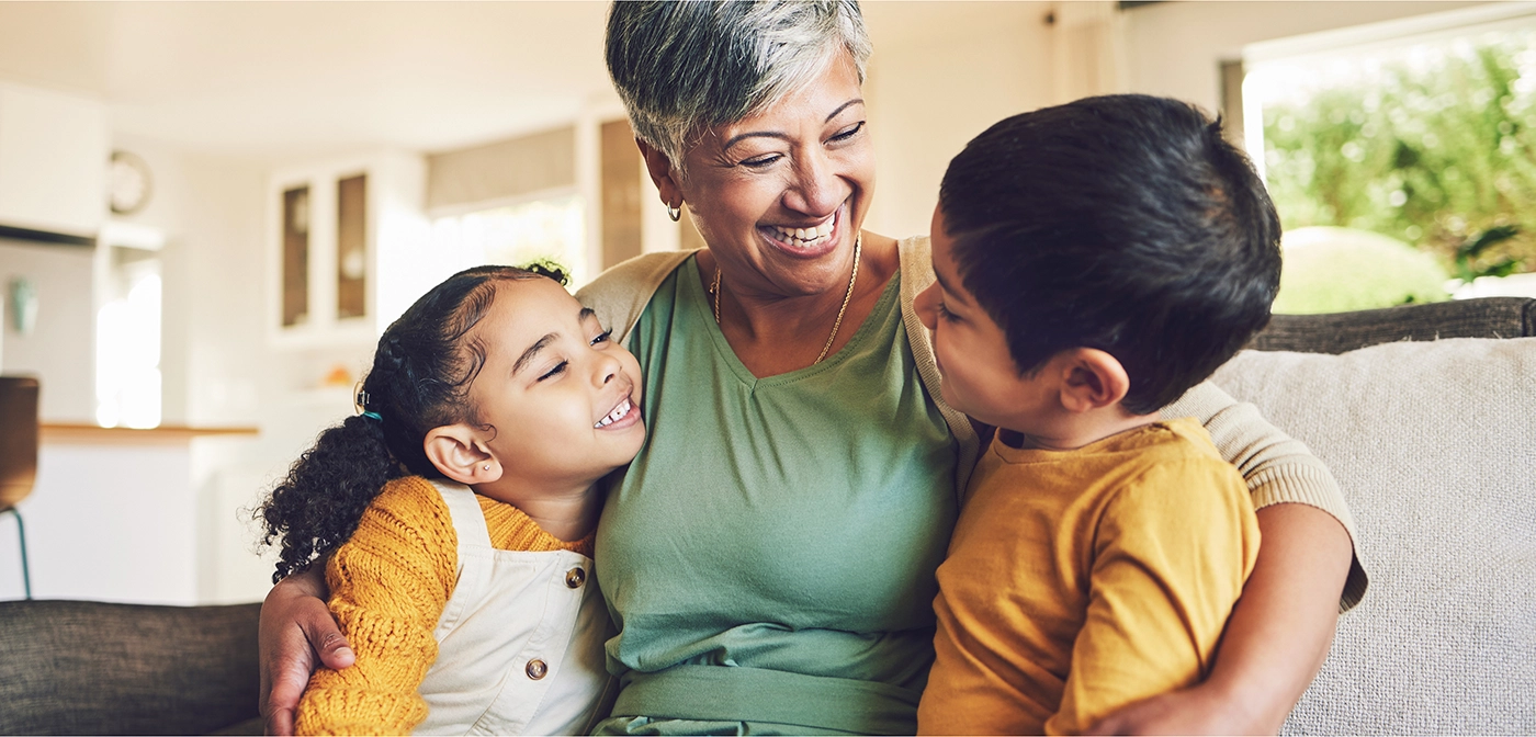 A light-medium skin woman sitting on a couch smiling and hugging two young children. Boy and girl are light-medium skin and smiling.