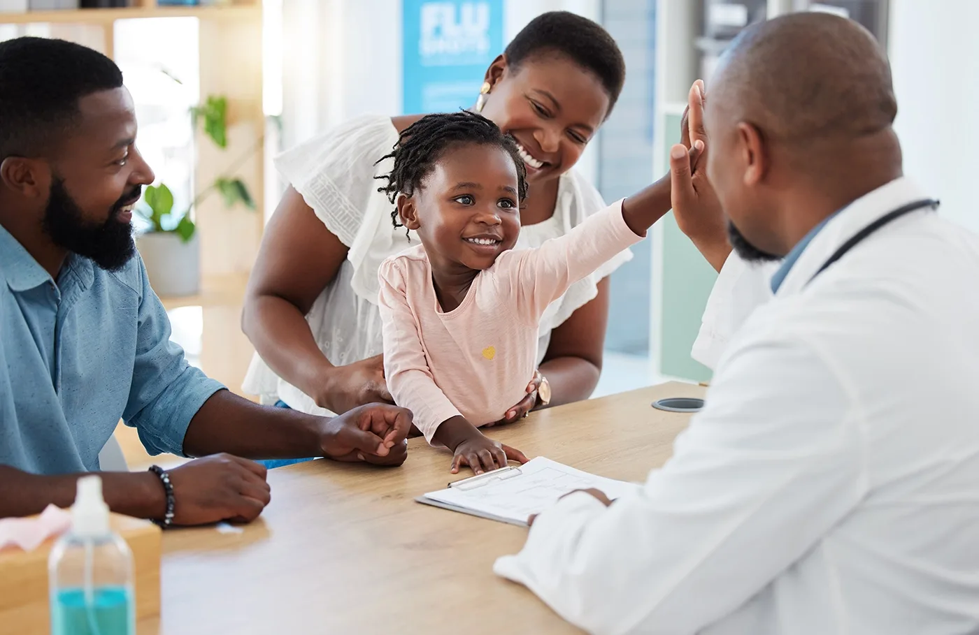 A young child is reaching out to a doctor across the table. Two adults are present with the child.