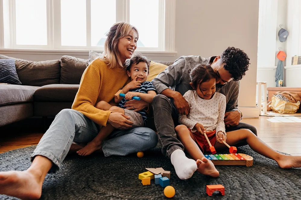 A family with medium-light skin tone sit on the living room floor, parents hugging their two children, while playing with a toy xylophone.