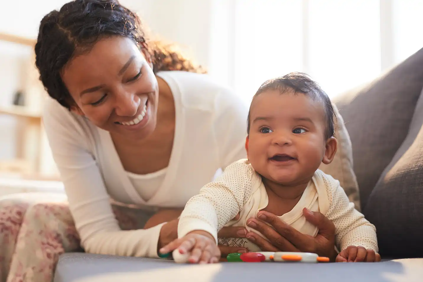 A woman with medium skin tone holds a baby with medium skin tone on a couch. The baby reaches for a colorful toy. Sunlight shines through a window.