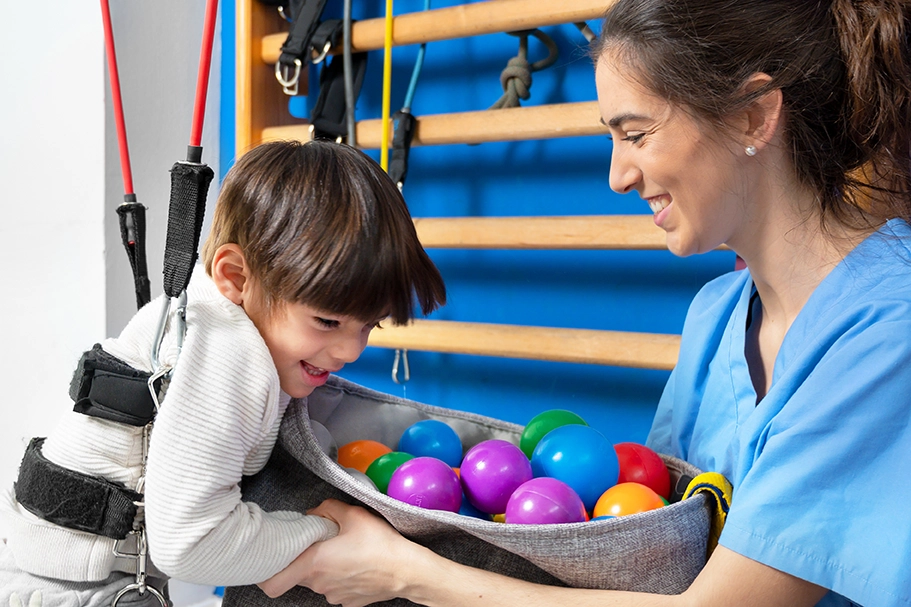 A smiling child with light skin tone is supported by a harness while reaching into a basket of colorful balls held by a person in a blue scrubs.
