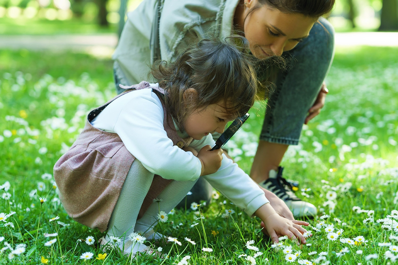A light skinned child crouches in a grassy field looking at flowers and an adult kneels nearby.